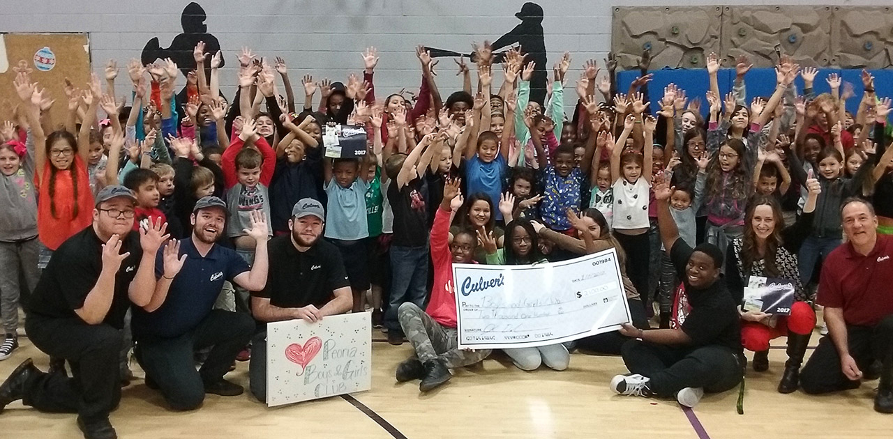 Group shot of Boy's and Girls Club kids and Culver's team members in gym holding hands up with giant Culver's check at center