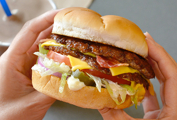 A Culver’s guest with a Bacon Deluxe in their hands as they prepare to take a bite. A Chocolate Shake is in the background on the table.